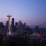 A panoramic view of a city skyline at dusk with the Space Needle and Mount Rainier in the background.