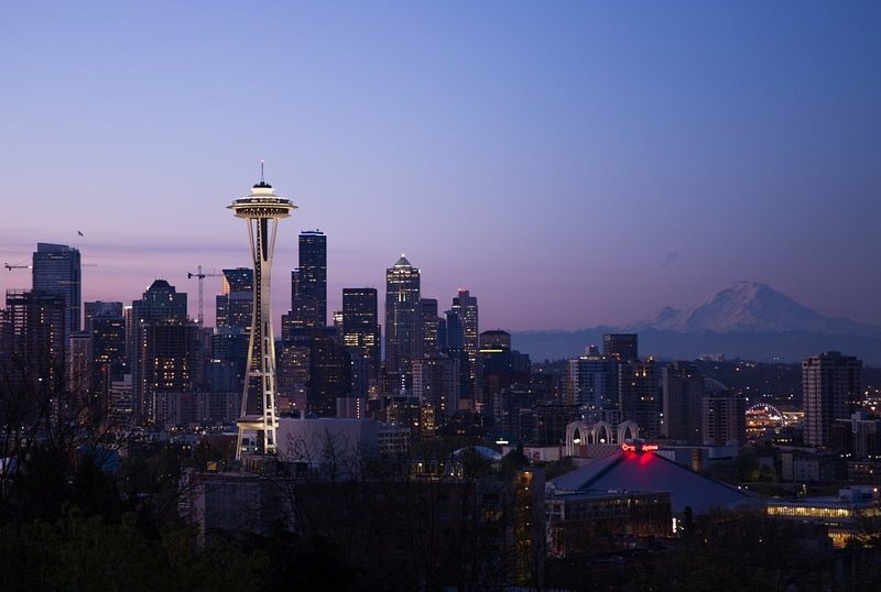 A panoramic view of a city skyline at dusk with the Space Needle and Mount Rainier in the background.