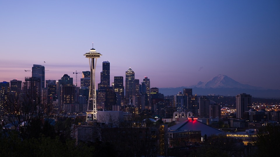 A panoramic view of a city skyline at dusk with the Space Needle and Mount Rainier in the background.